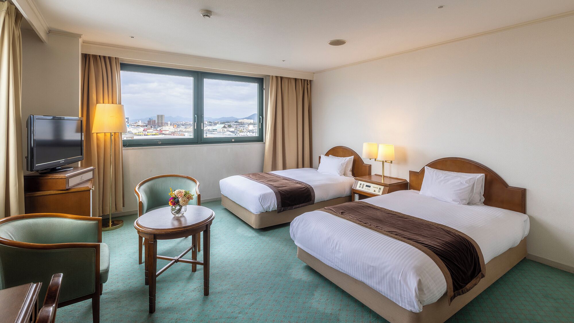 A hotel room with two beds, a TV, a desk, a chair, and a view of the city.