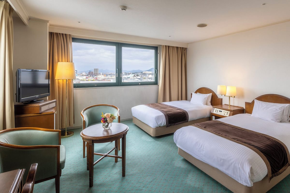 A hotel room with two beds, a TV, a desk, a chair, and a view of the city.