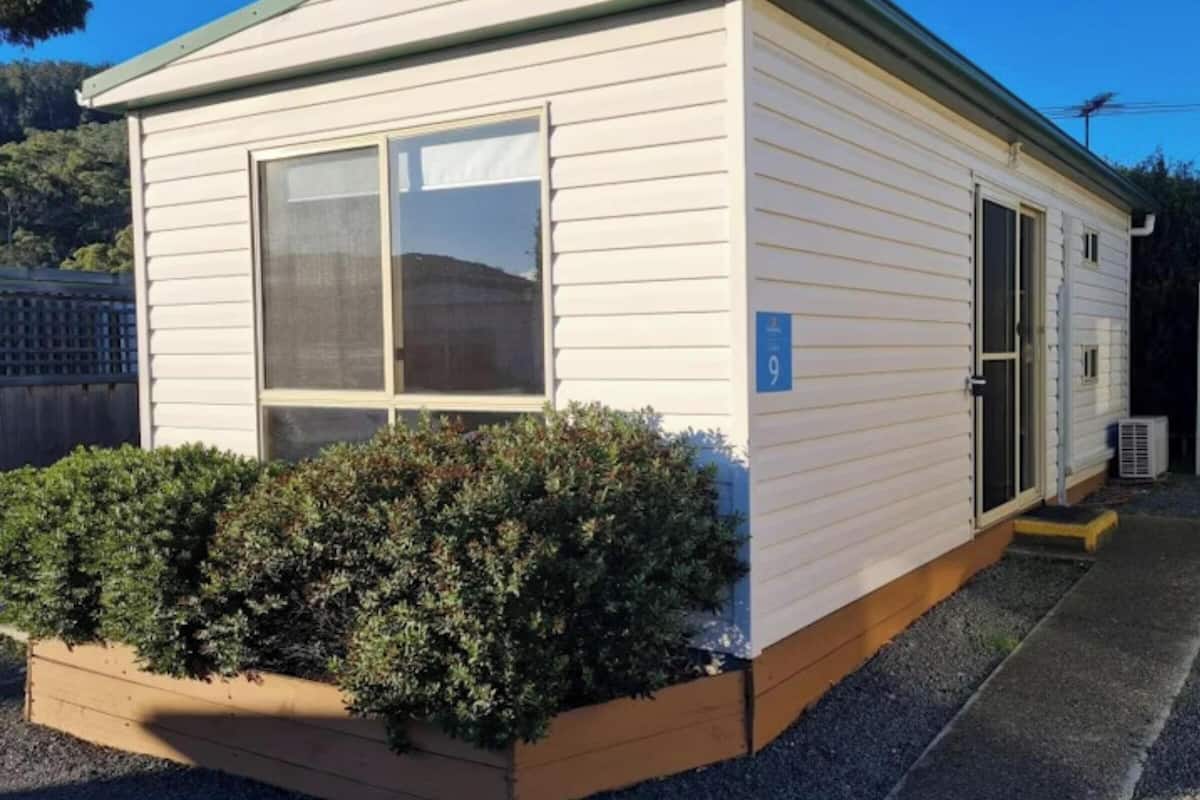 A small, single-story mobile home with a window and door, a number sign, and a small garden area.