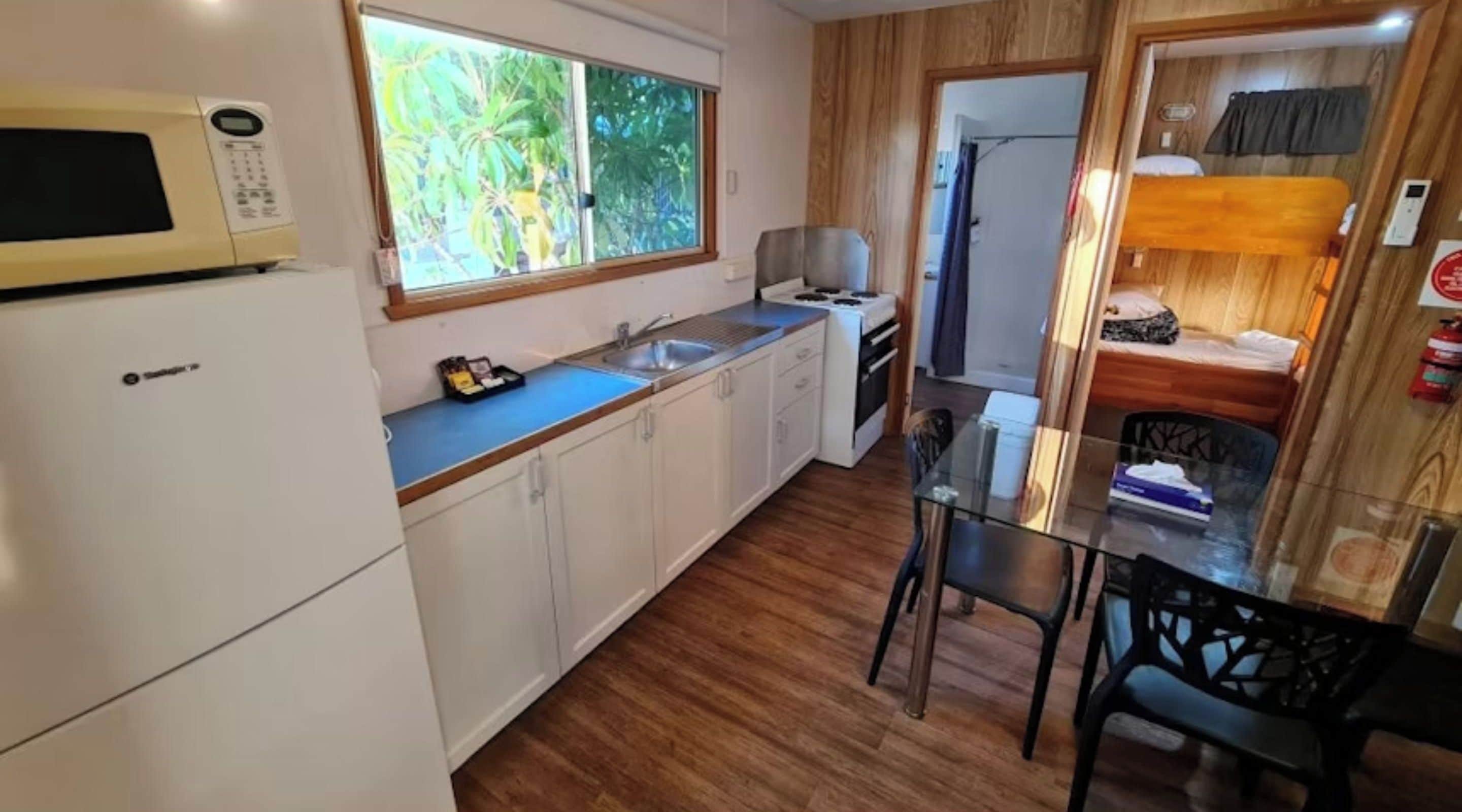 A compact kitchen with a microwave, sink, and stove, a dining area with a table and chairs, and a small sleeping area with a bed and window.