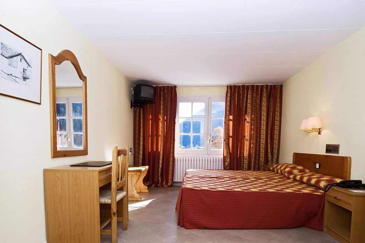 A bedroom with a bed, a desk, a chair, a mirror, a window with curtains, and a lamp.