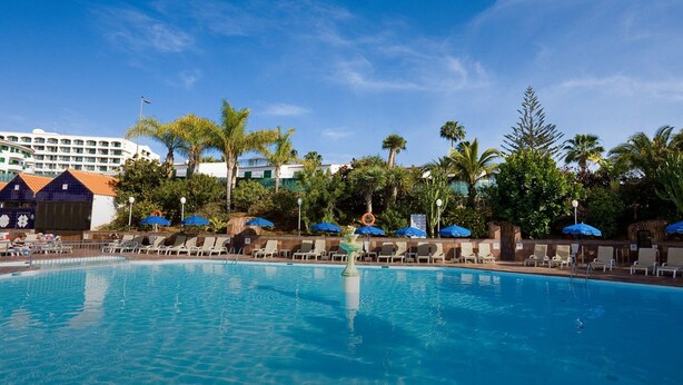 A swimming pool with lounge chairs and umbrellas, surrounded by palm trees and a hotel building in the background. - Residencia Daina (Arinsal)