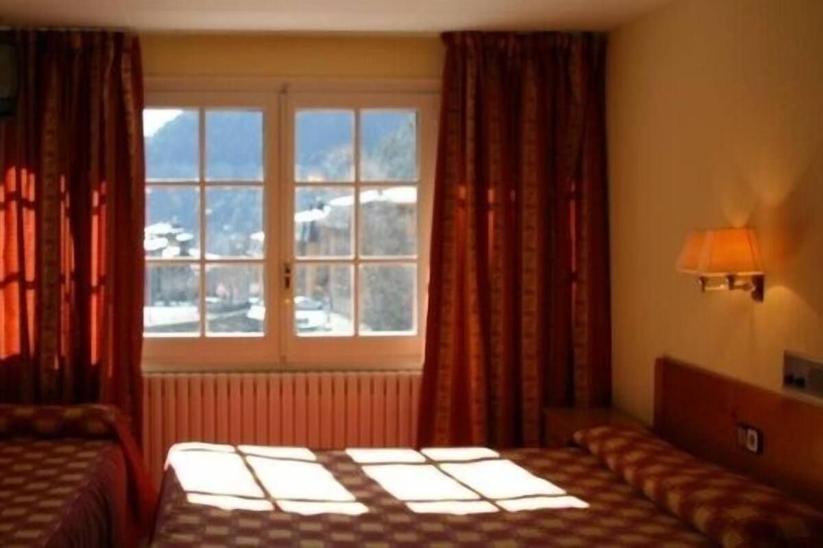 A hotel room with two beds, a window with a view of mountains, a radiator, and a wall-mounted lamp.