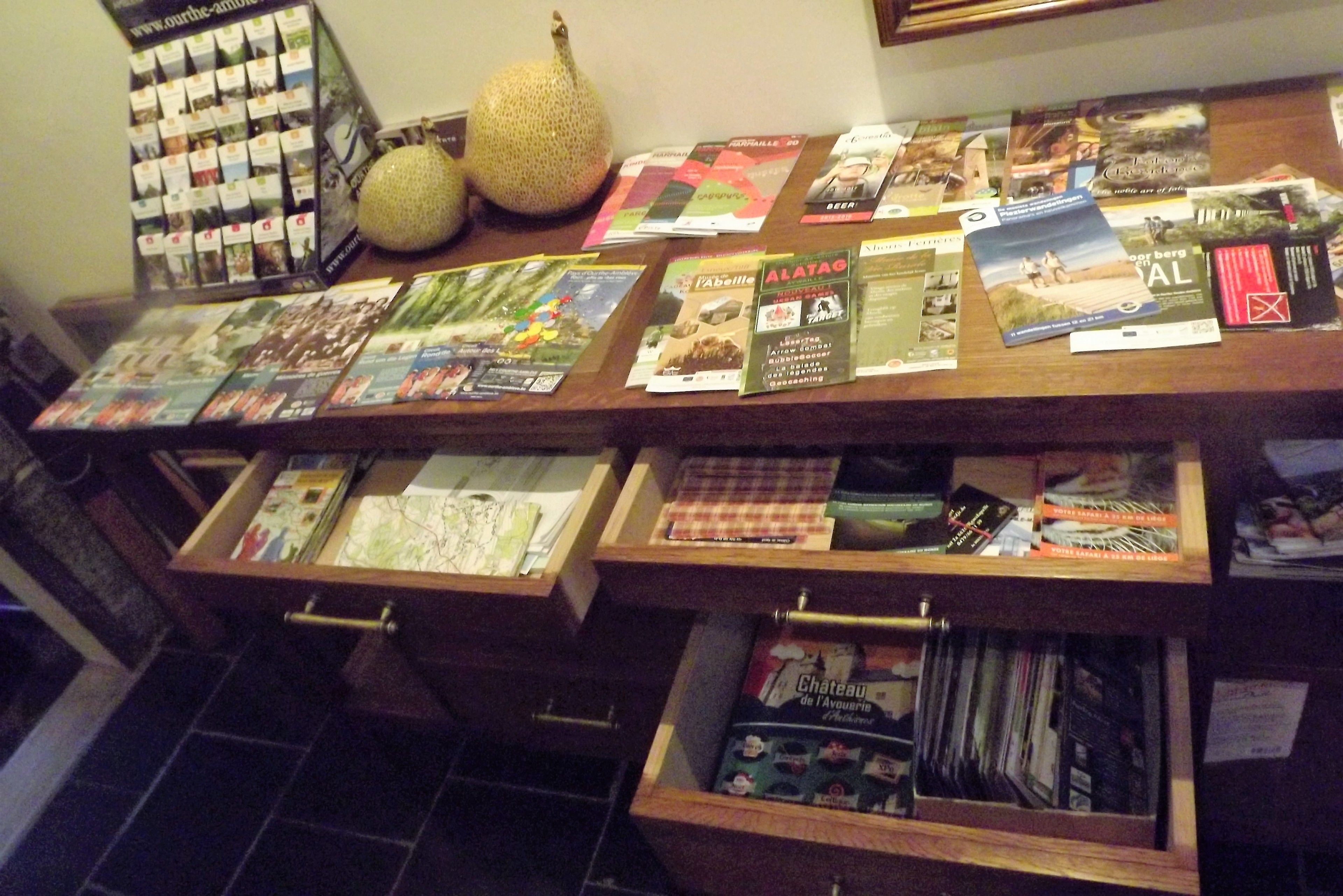 Une table d’exposition en bois, sur laquelle sont disposés divers dépliants et magazines, un panier de fruits et un trousseau de clés.