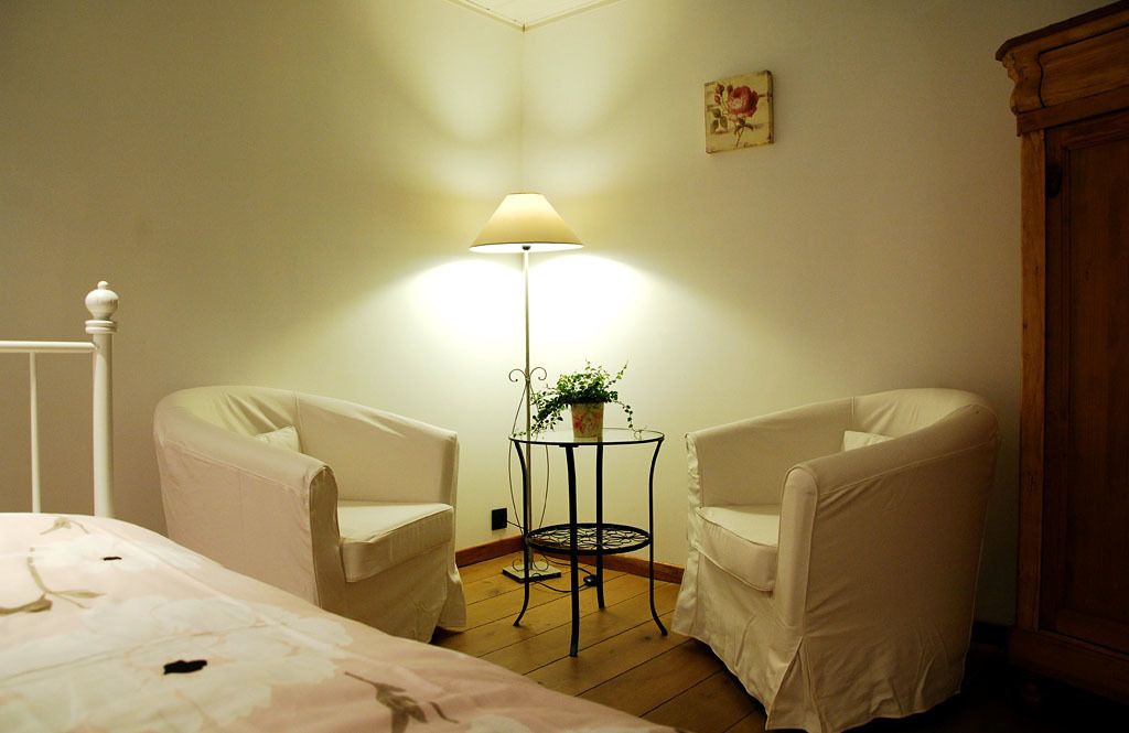 Una camera da letto con un letto, due poltrone bianche, un tavolino con una lampada e un armadietto in legno.