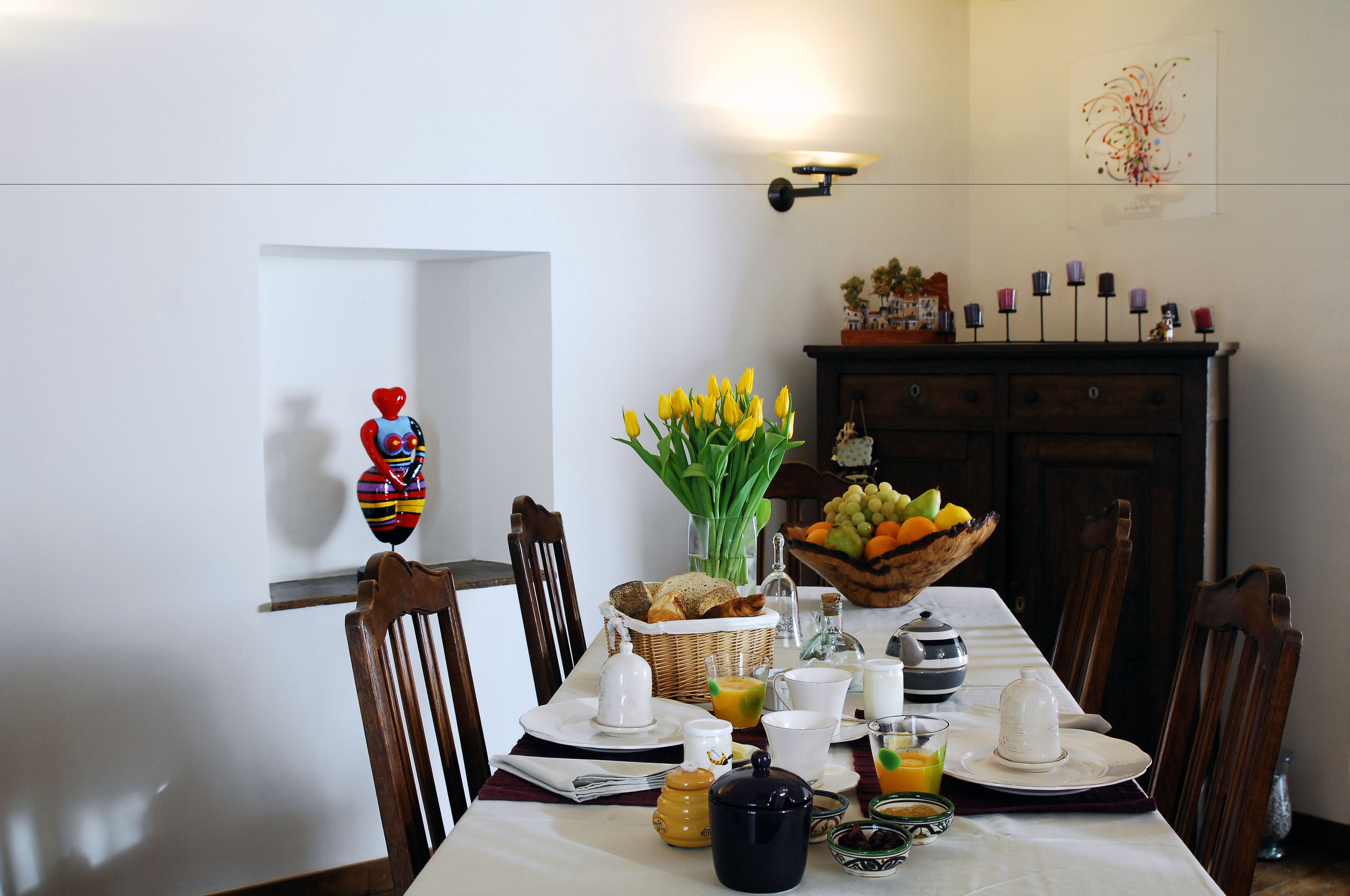 A dining table set with breakfast items, a vase of yellow flowers, and a fruit bowl.