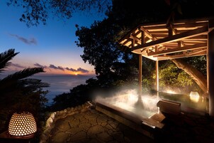 A hot spring area with a wooden pergola, stone flooring, and a view of the sunset over the ocean.