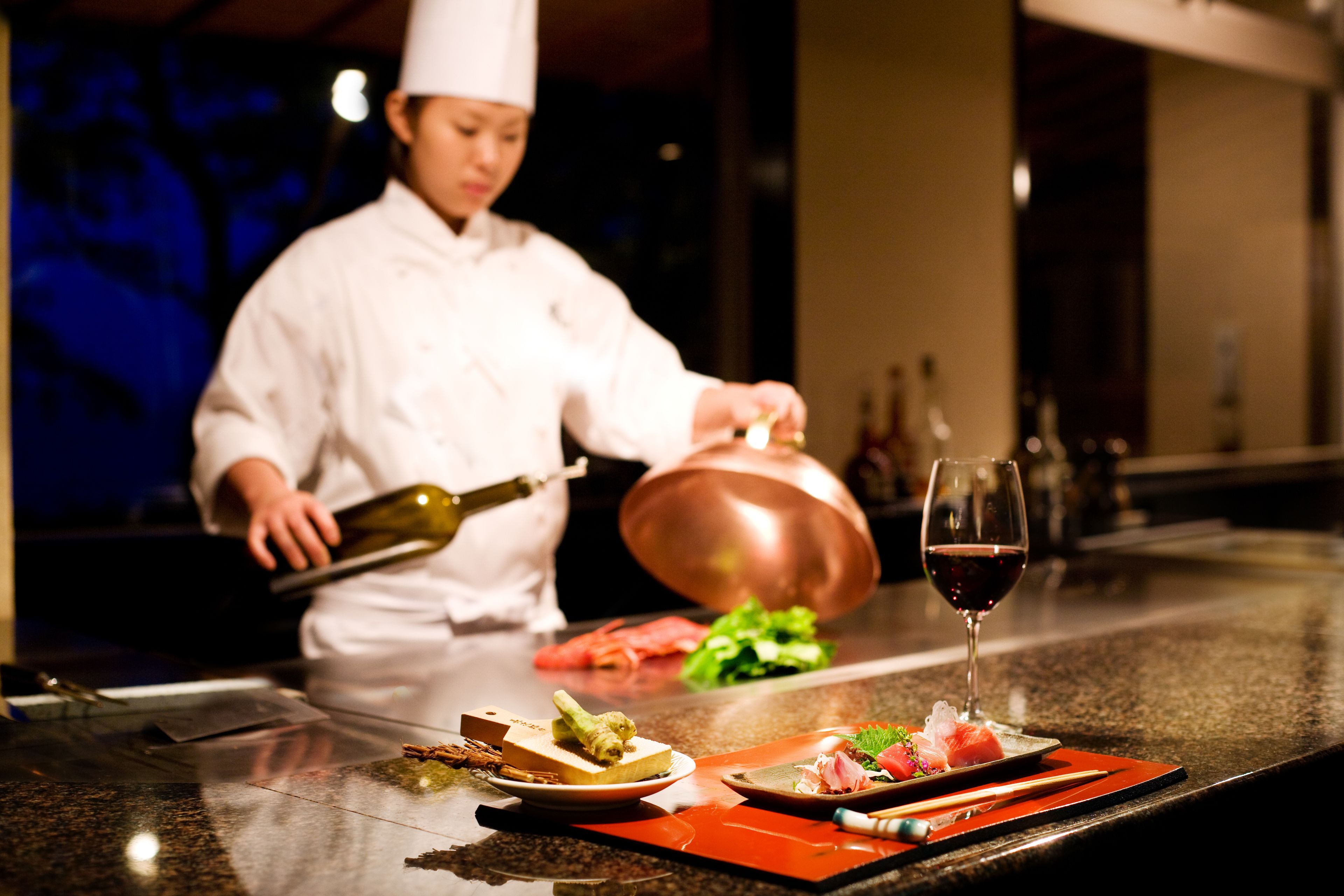 A chef preparing food in a kitchen with a copper pot and a wine glass on the counter.