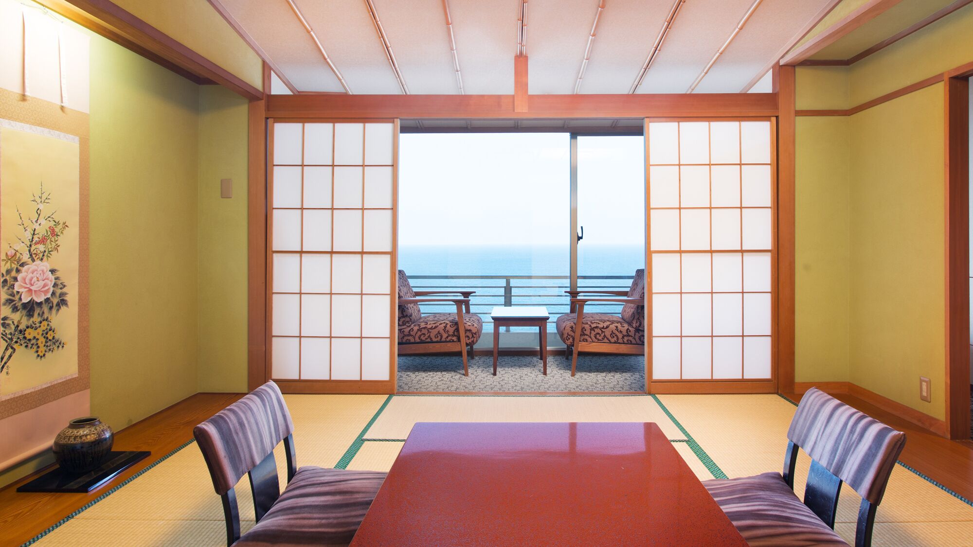 A traditional Japanese room with tatami flooring, a low table, two chairs, and a sliding door leading to a balcony with a view of the sea.