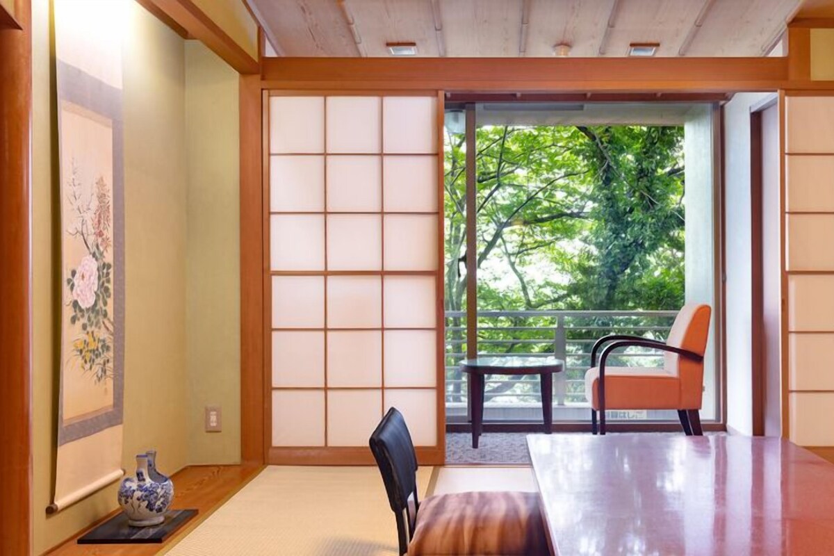 A traditional Japanese room with tatami flooring, a low table, and a sliding door leading to a balcony with a view of trees.