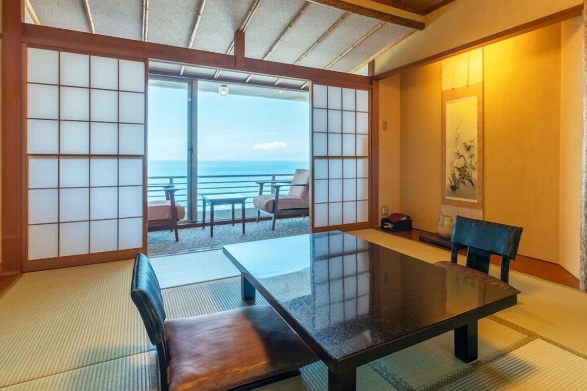 A traditional Japanese room with tatami flooring, a low table, and sliding doors leading to a balcony with a view of the sea.