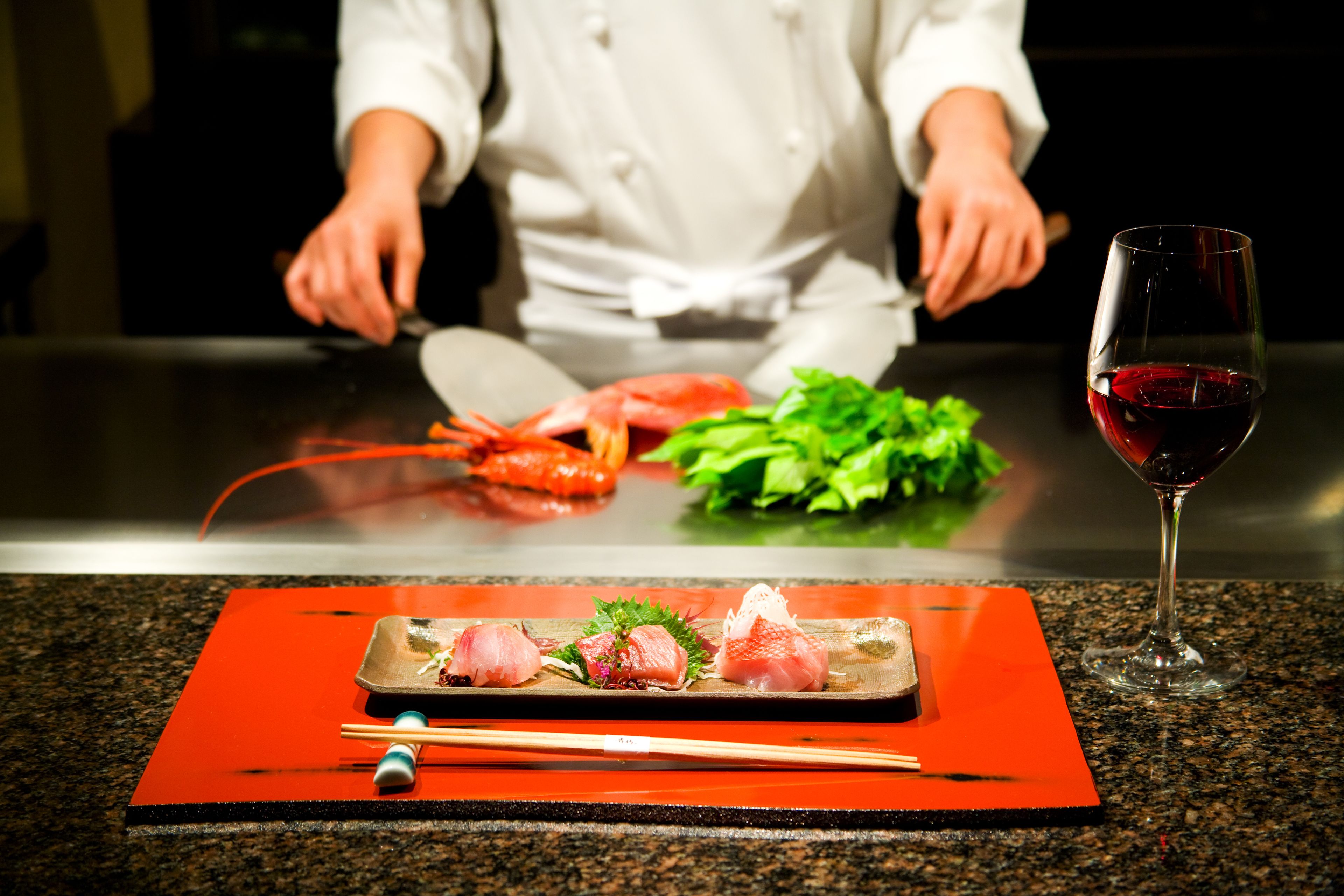A chef preparing food at a counter with a plate of sashimi and a glass of red wine.