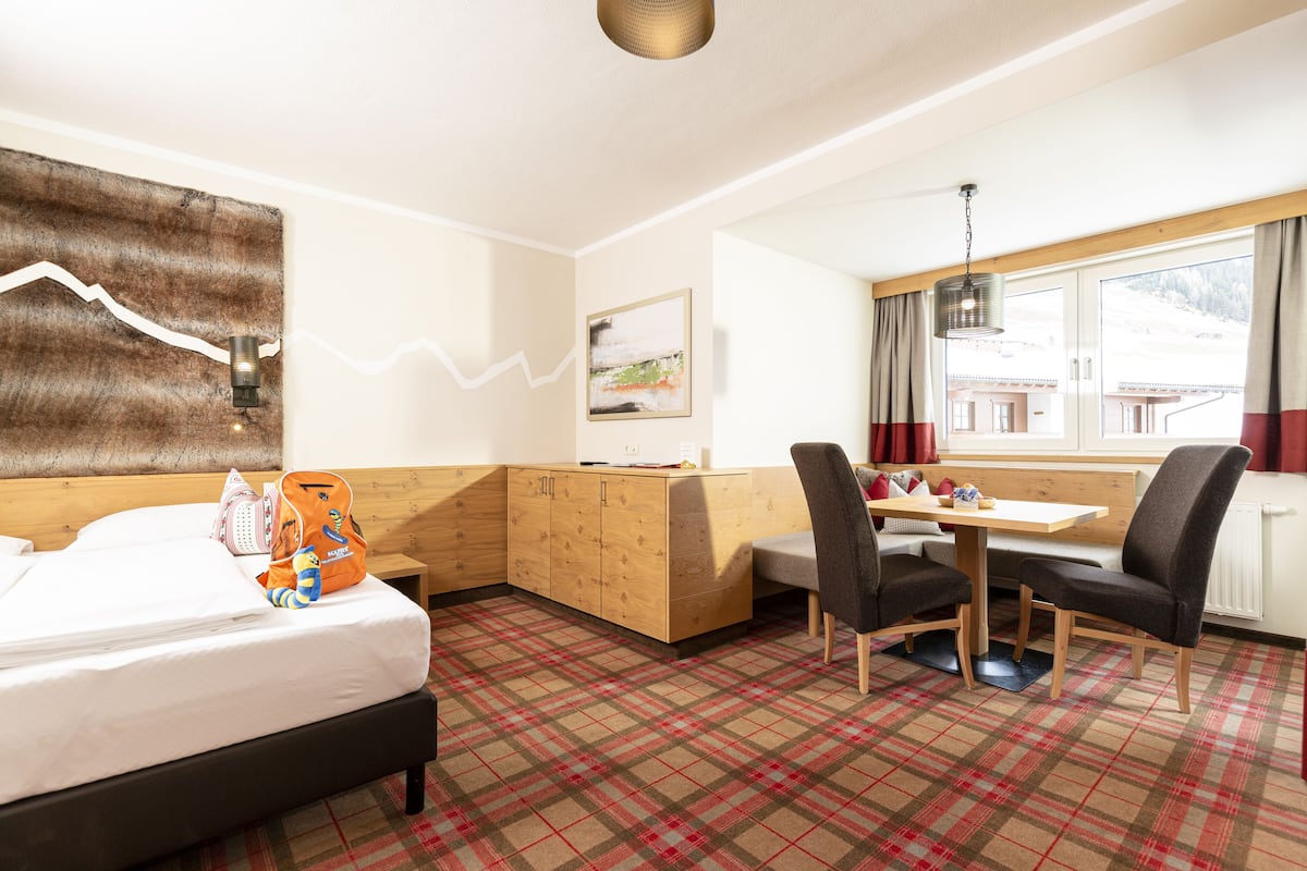 Habitación de hotel con cama, un banco de madera, una mesa de comedor con sillas y vista a un paisaje nevado a través de la ventana.