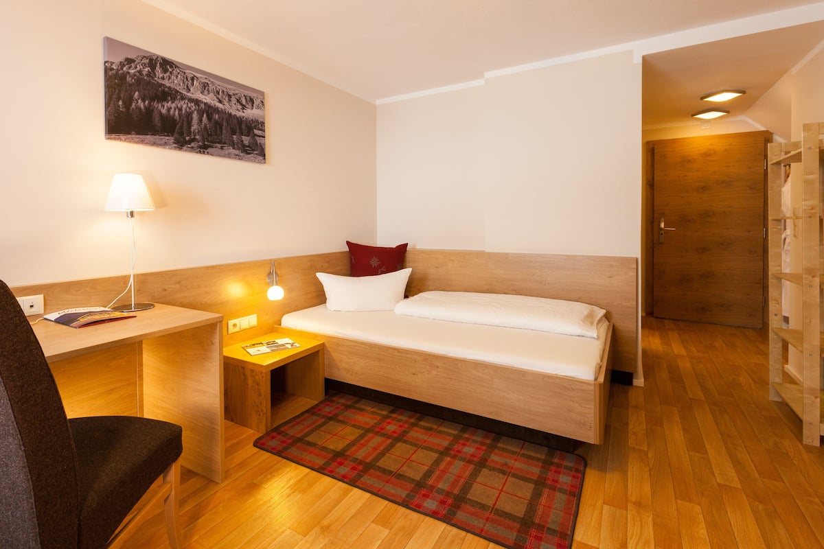 Una habitación de hotel compacta con una cama de madera, un escritorio con lámpara, una silla y un tapete a cuadros.