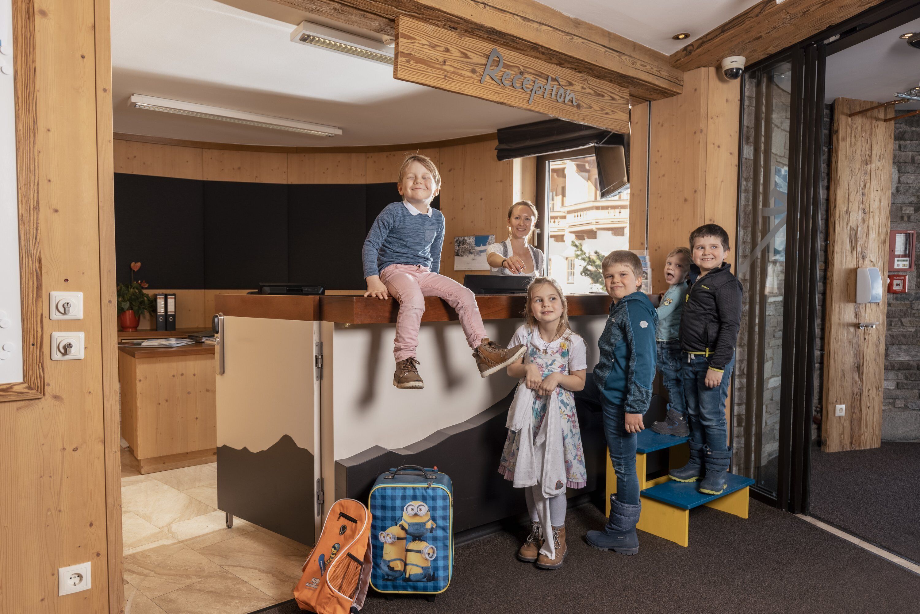 A reception area with wooden walls, a reception desk, and a sign labeled "Reception." There are children standing and sitting around the desk, and a suitcase on the floor.