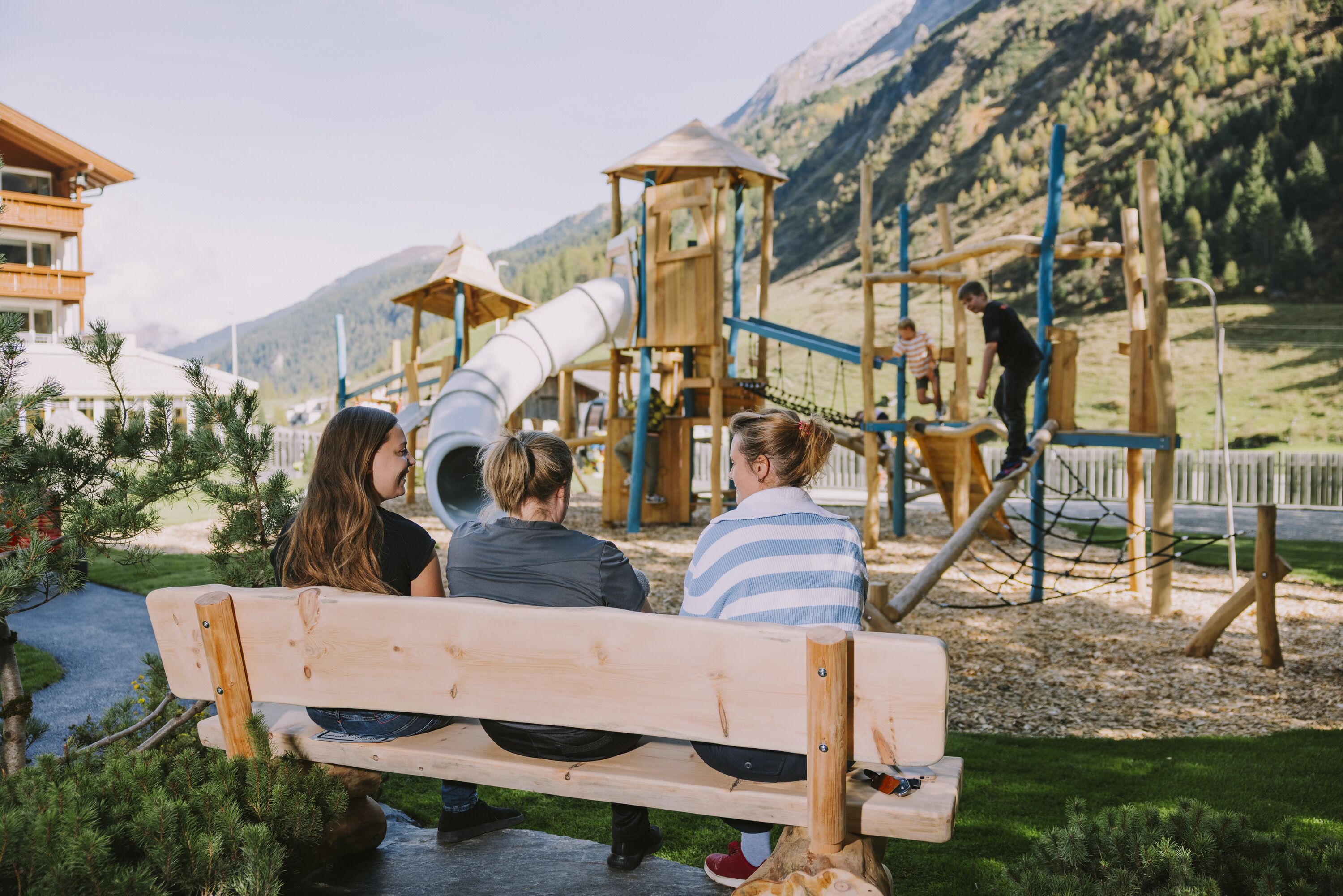 A wooden bench in a park with a playground and mountains in the background.