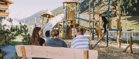 A wooden bench in a park with a playground and mountains in the background.