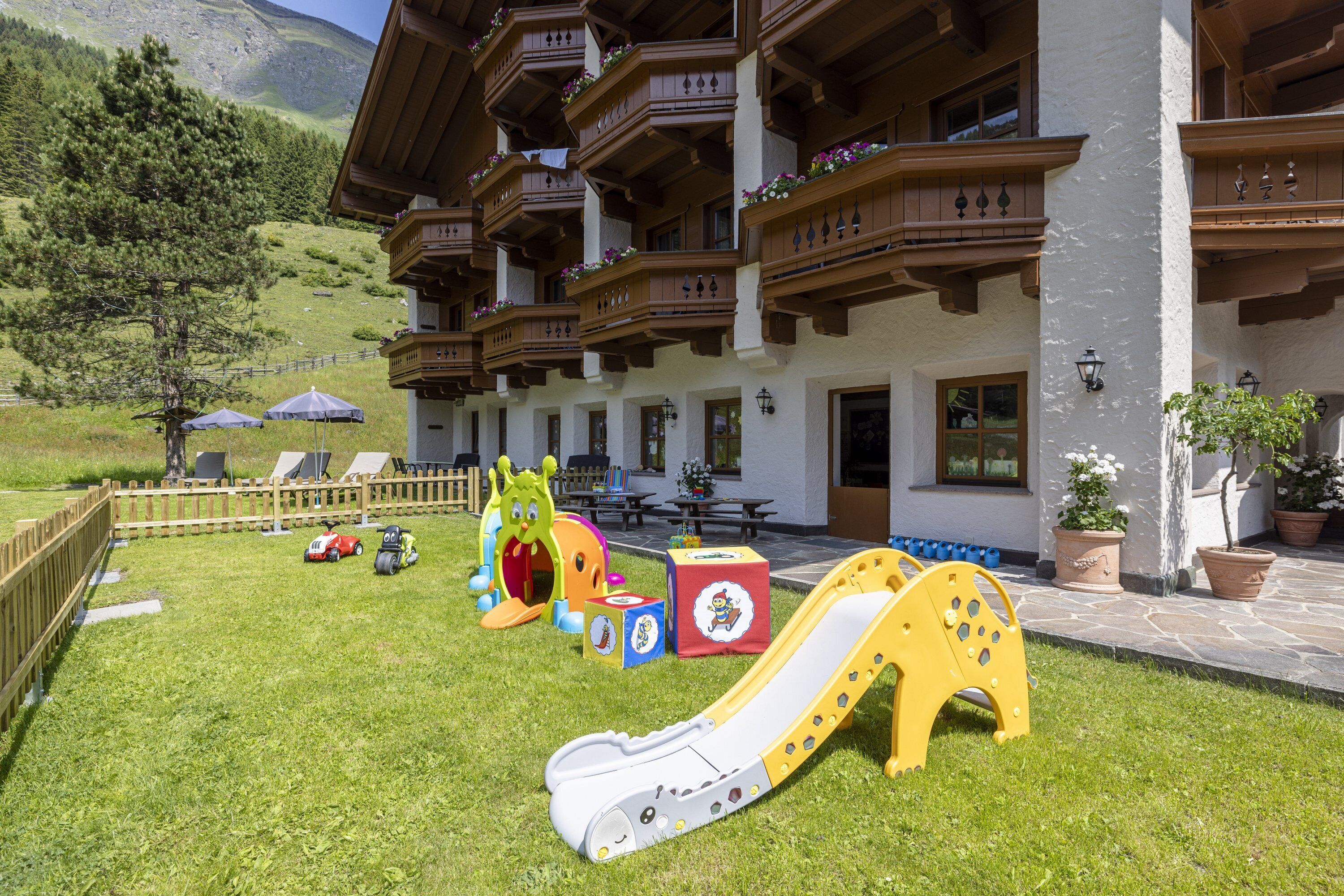 A children's play area with a slide, toys, and a picnic table in front of a hotel with balconies and a mountain view.