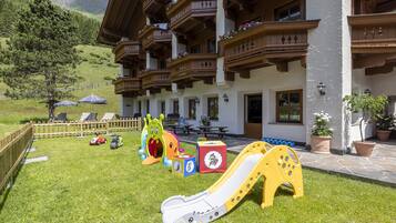 A children's play area with a slide, toys, and a picnic table in front of a hotel with balconies and a mountain view.