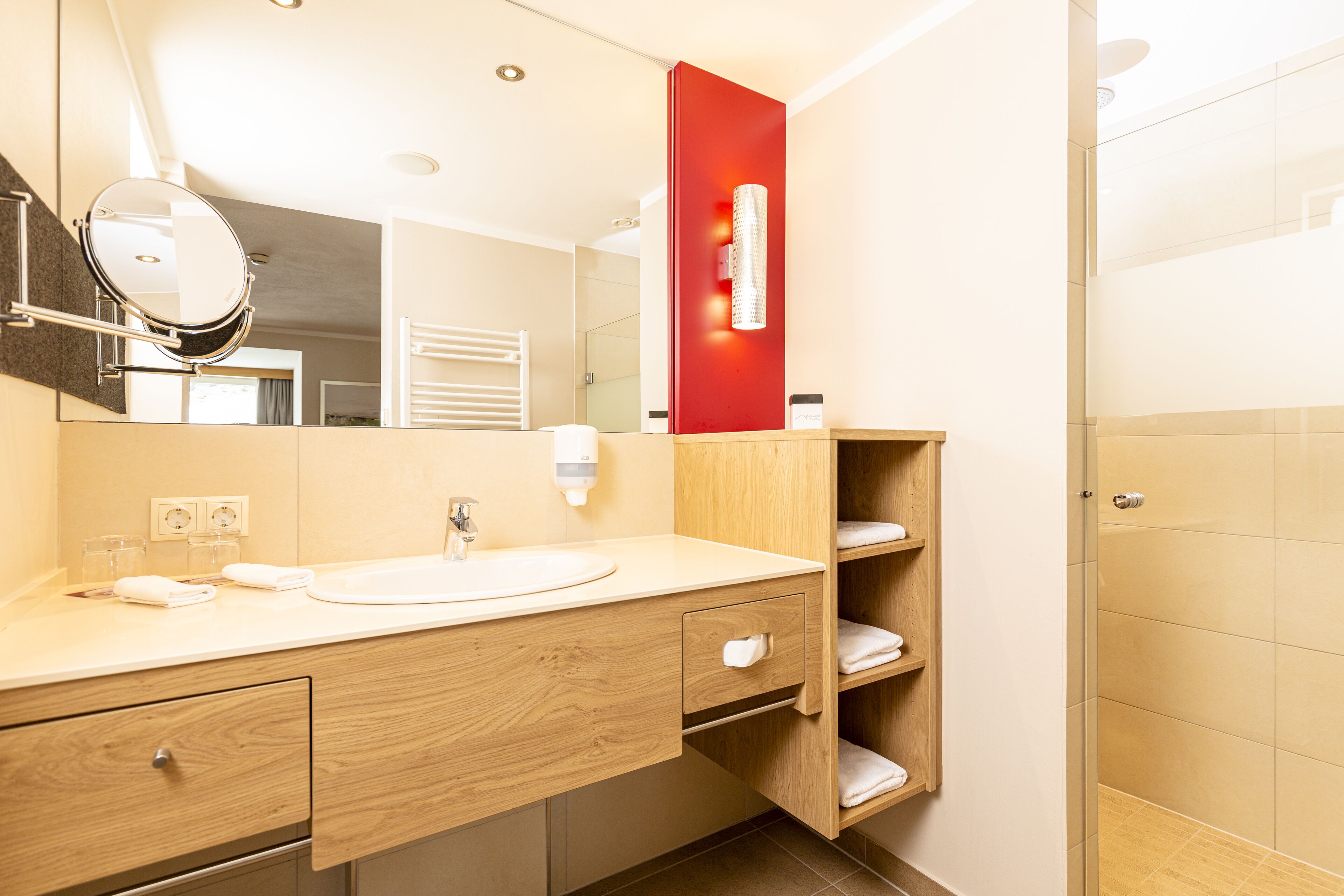 A modern bathroom with a wooden vanity, a round mirror, a red accent wall, and a glass shower enclosure.