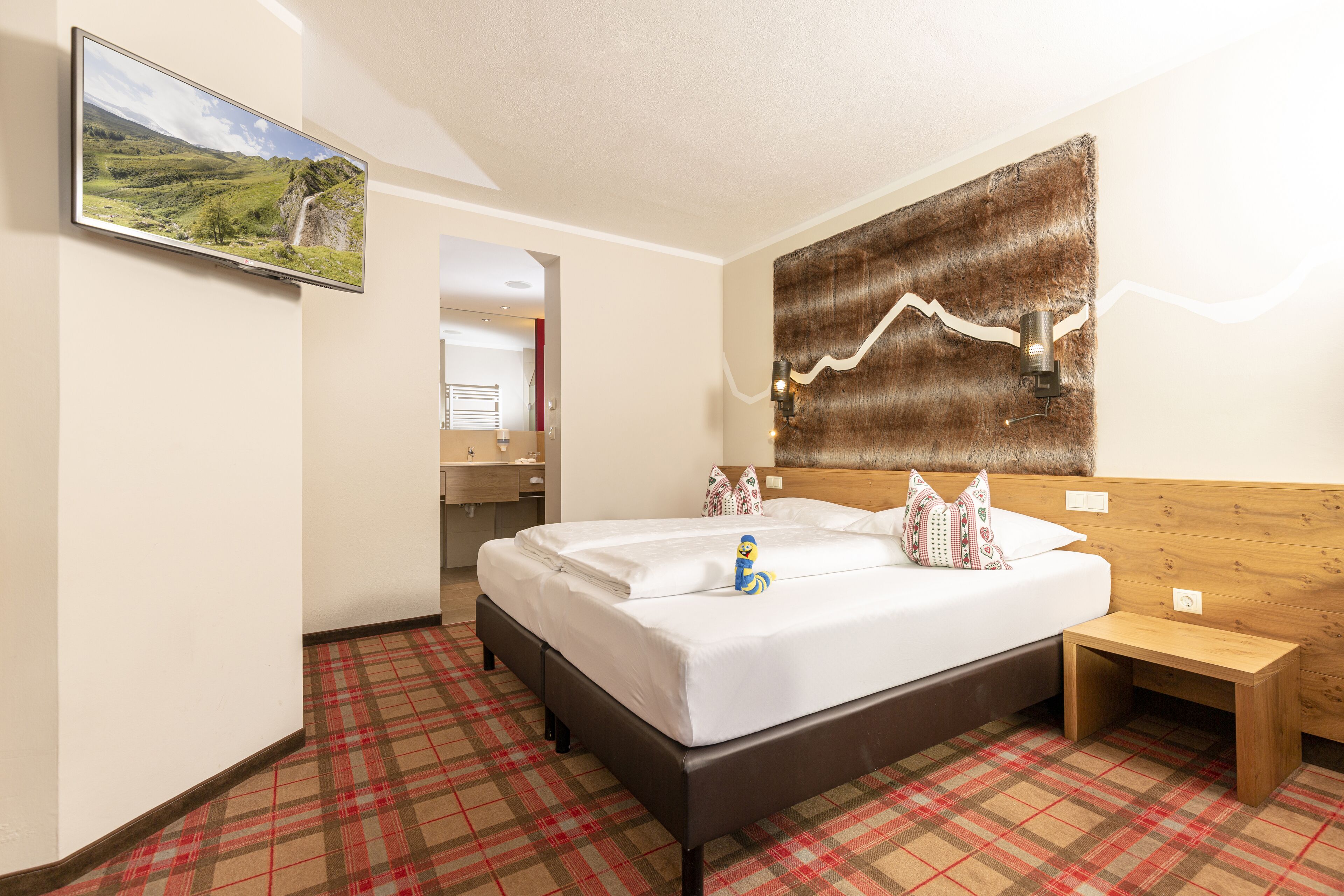 A hotel room with a bed, a wooden headboard, a small bedside table, a wall-mounted TV, and a plaid-patterned floor.