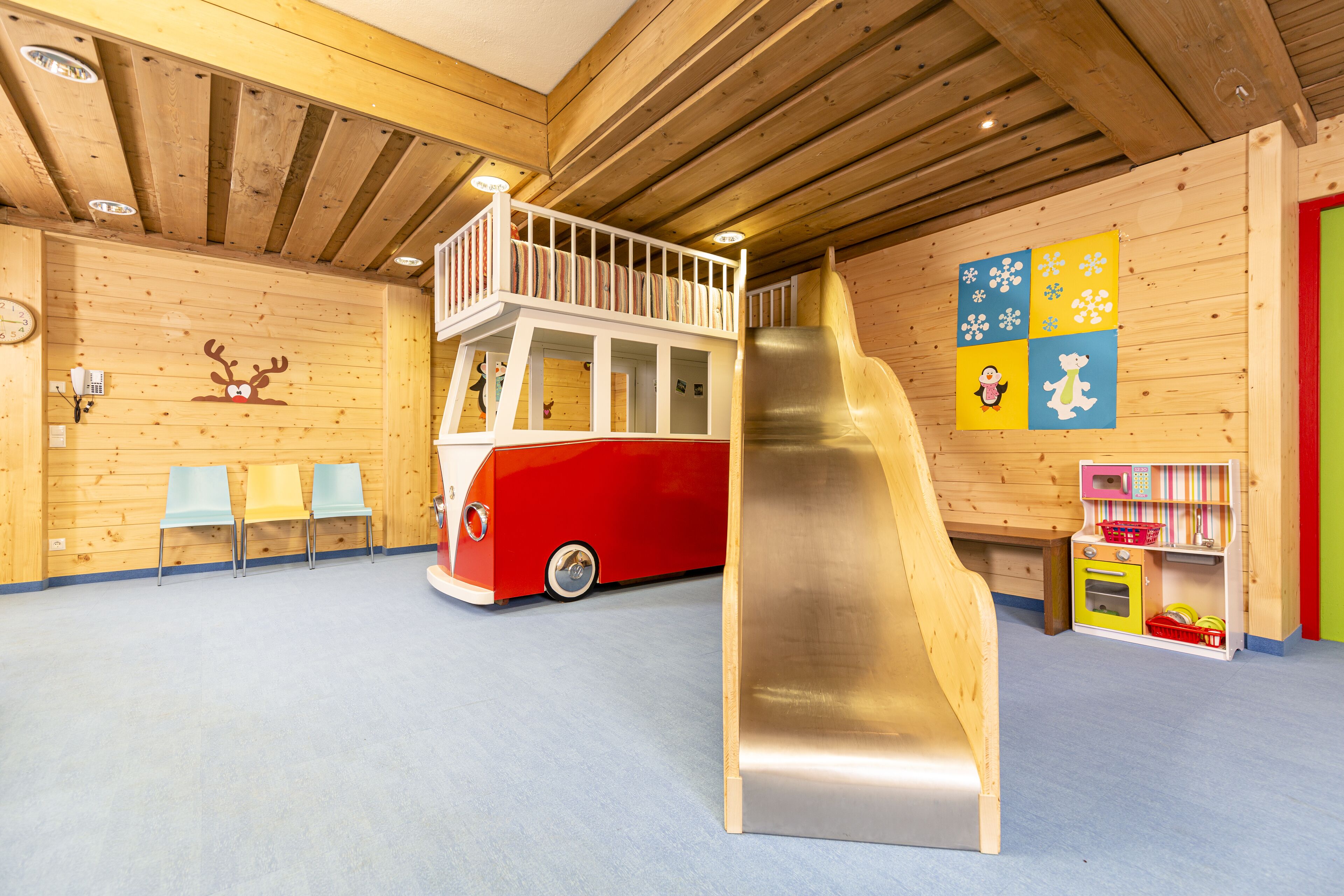 Zona de juegos infantiles con un tobogán, una casita con forma de autobús rojo y detalles de madera.
