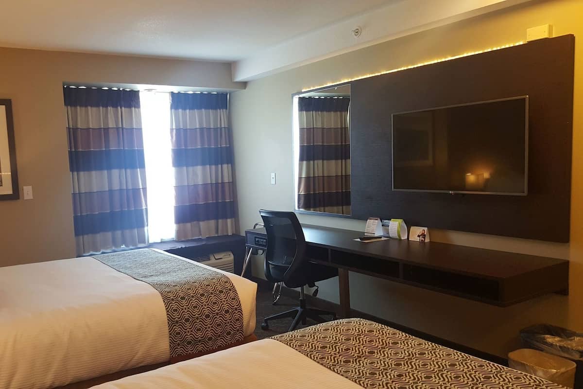Ein Hotelzimmer mit zwei Betten, einem Schreibtisch, einem Sessel, einem Fernseher und einem gerahmten Bild an der Wand.