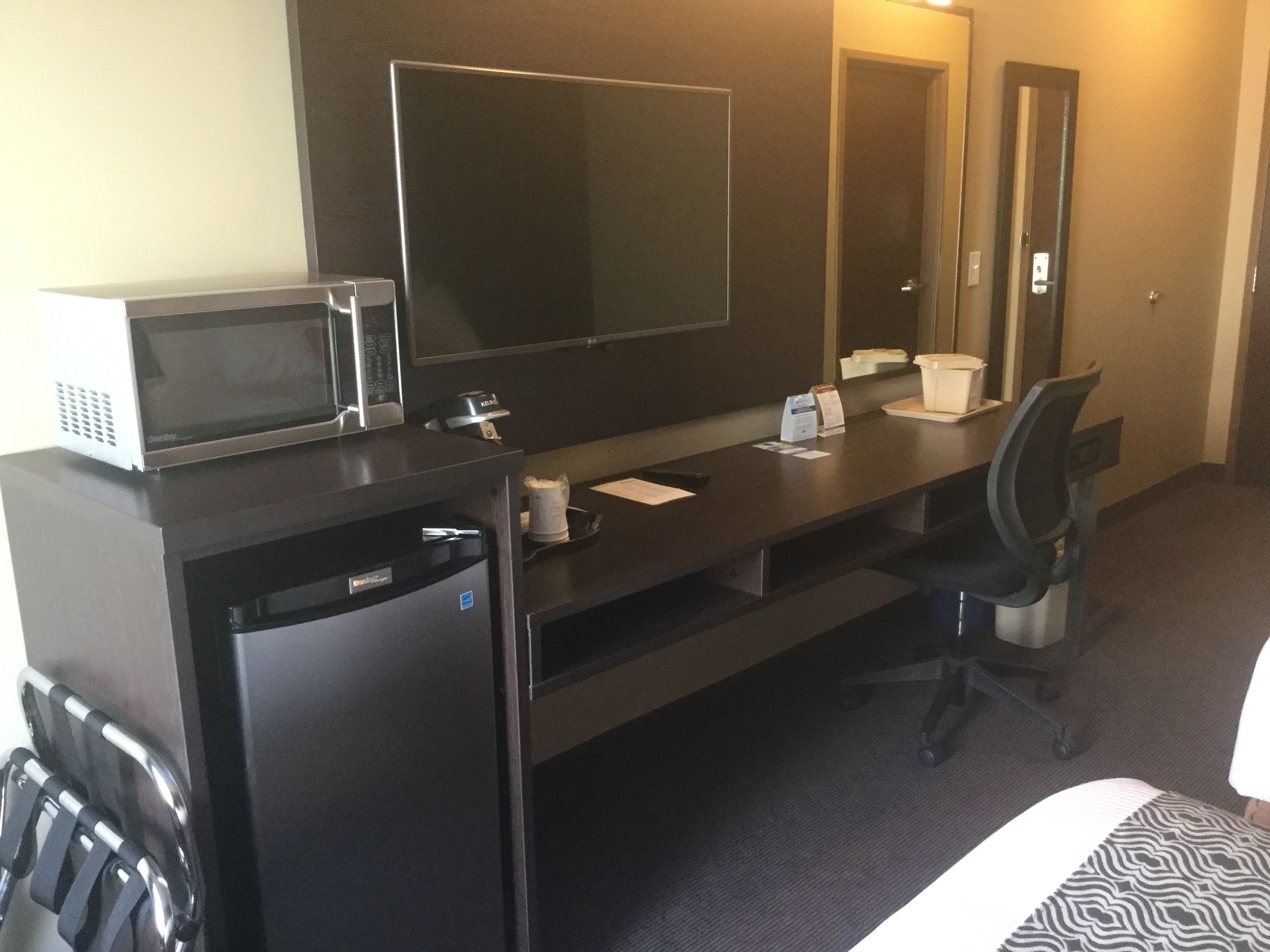 A hotel room with a dark wooden desk, a microwave, a refrigerator, a chair, and a large mirror.