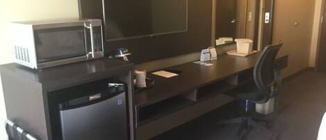 A hotel room with a dark wooden desk, a microwave, a refrigerator, a chair, and a large mirror.