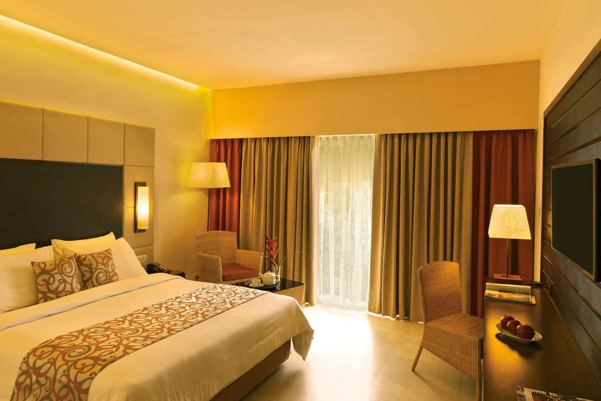 Une chambre d’hôtel avec un grand lit, un coin salon avec un fauteuil, une télévision et un bureau avec une lampe.
