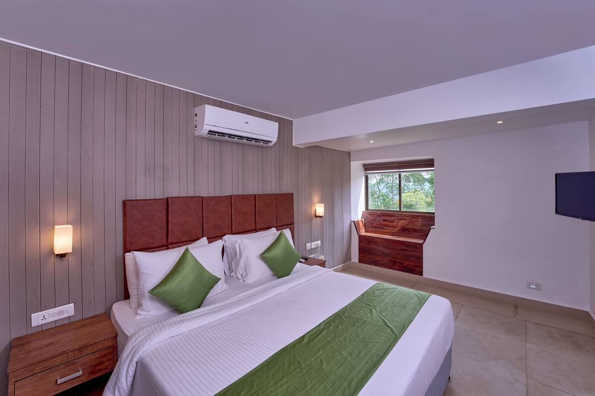 Une chambre d’hôtel moderne avec un grand lit, une table de chevet, une tête de lit en bois, un climatiseur mural, une télévision et une fenêtre donnant sur un paysage verdoyant.