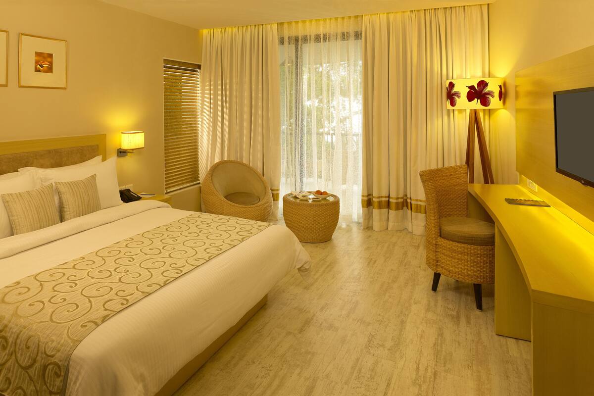 Une chambre d’hôtel avec un grand lit, un bureau avec une télévision, une chaise et une petite table ronde avec des fruits.