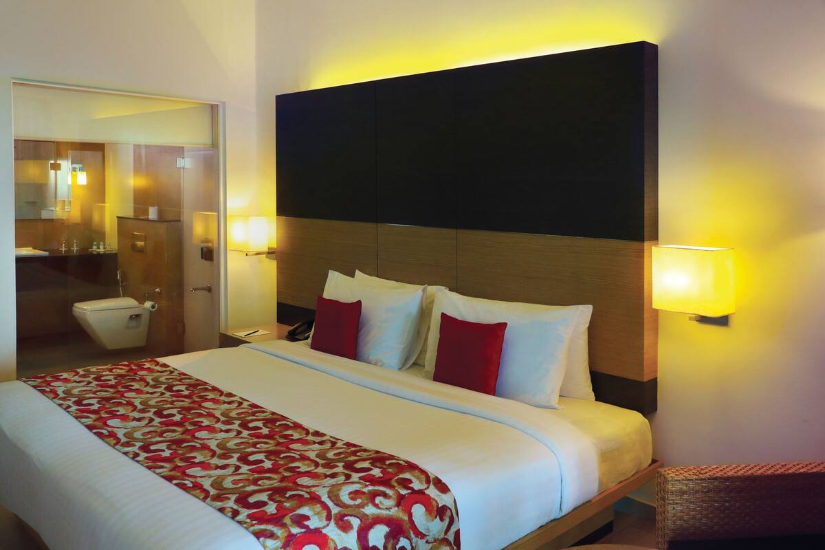 Une chambre d’hôtel avec un grand lit, une table de chevet, une commode, un miroir et une salle de bain visible à travers une porte ouverte.
