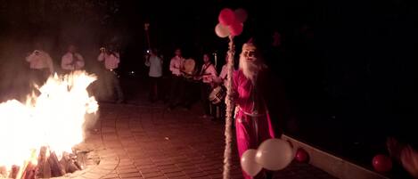 Una persona vestida de rosa sostiene globos en un evento nocturno, con un fuego de fondo.