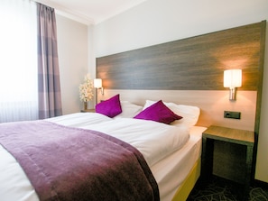 Quarto de hotel com cama grande, duas almofadas, mesa de cabeceira, abajur e um vaso com flores.
