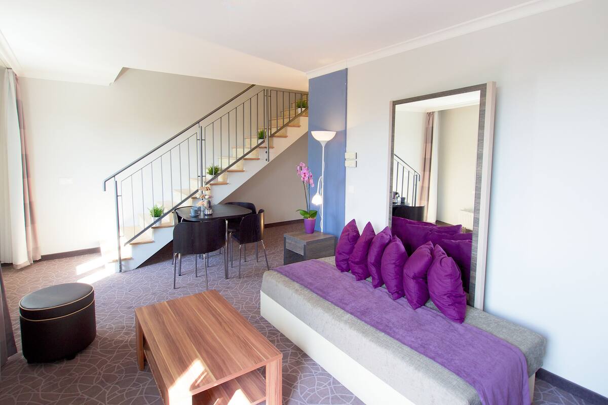 Ein modernes Wohnzimmer mit einer Couch, einem Couchtisch und Sesseln. Eine Treppe mit Geländer und Topfpflanzen ist ebenfalls zu sehen.