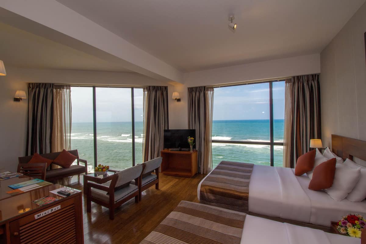A hotel room with a large bed, a sofa, a desk with a computer, and a TV. The room has large windows with ocean views.