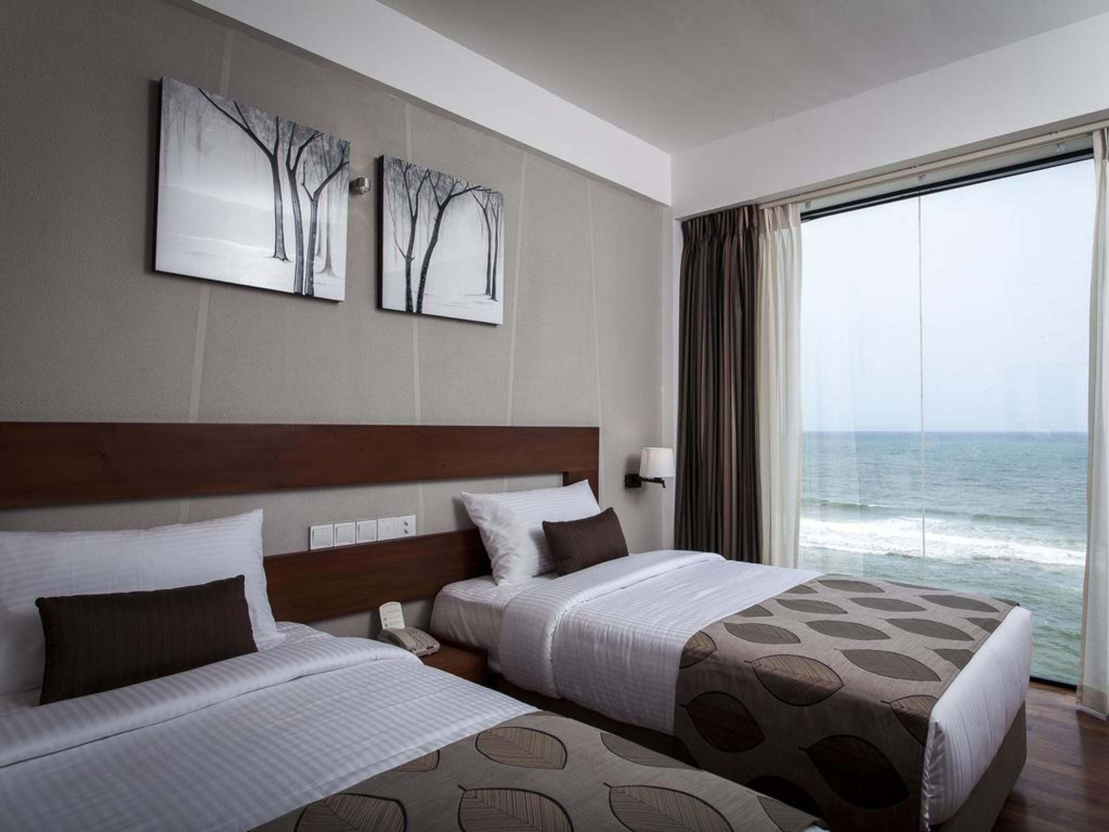 Une chambre d’hôtel avec deux lits, une tête de lit en bois, une table de chevet avec un téléphone et une vue sur l’océan grâce à de grandes fenêtres.
