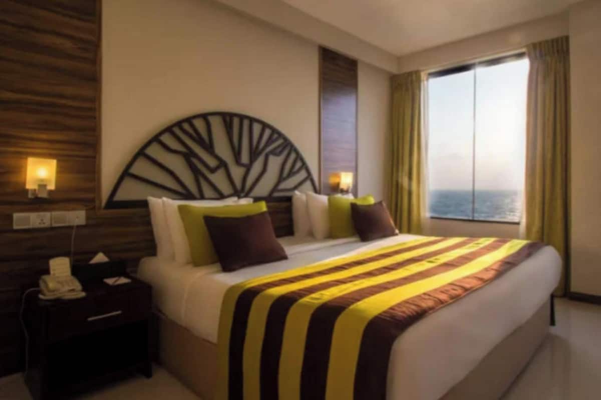 A hotel room with a large bed, a nightstand, a wall-mounted light, and a view of the sea through a large window.