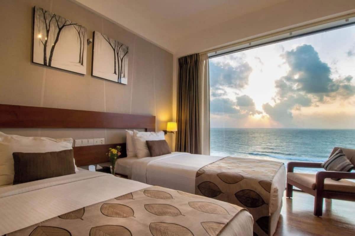 A hotel room with two beds, a large window with a view of the sea, and artwork on the wall.