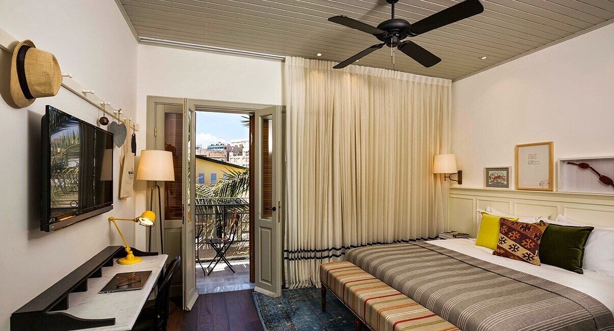 A hotel room with a bed, bedside lamps, a desk with a lamp, a ceiling fan, a TV, and a view through a door.