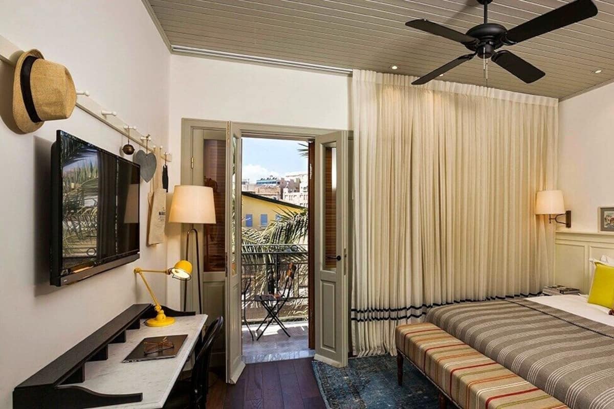 Une chambre d’hôtel avec un lit, des lampes de chevet, un bureau avec une lampe, un ventilateur de plafond, une télévision et une vue sur l’extérieur, visible à travers une porte.