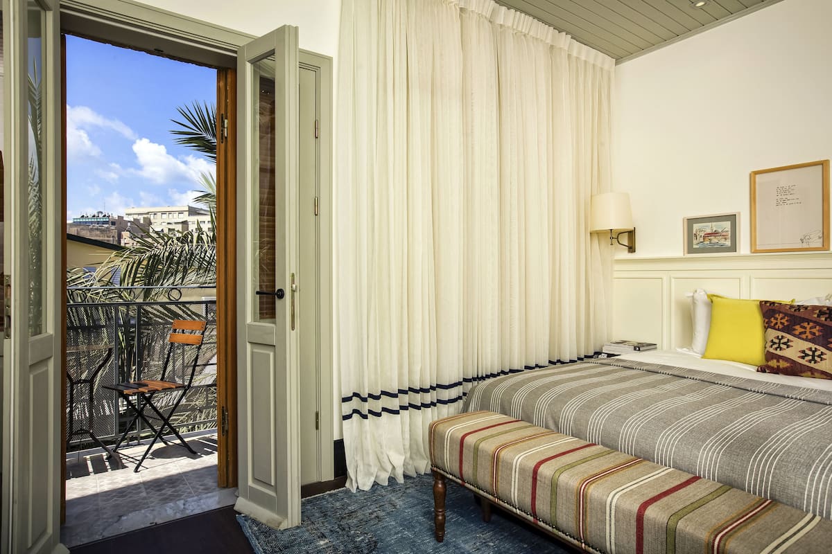 Une chambre d’hôtel avec un lit, une banquette, une chaise, une fenêtre avec des rideaux, une lampe et un tableau encadré au mur.