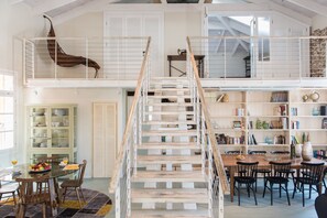 Een ruime loft met een houten trap, een eetgedeelte met een glazen tafel en een bibliotheek met boekenkasten.