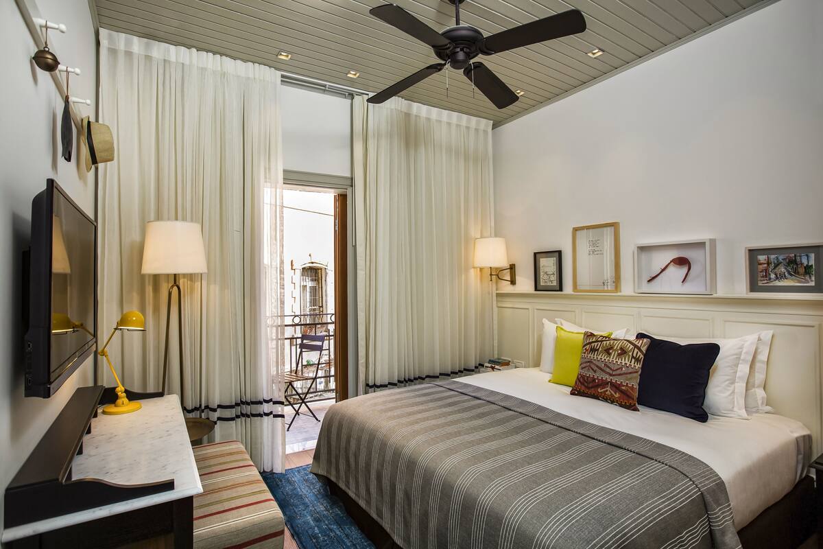 Une chambre d’hôtel avec un lit, un bureau avec une lampe, un ventilateur de plafond et une vue sur l’extérieur par une fenêtre munie de rideaux.