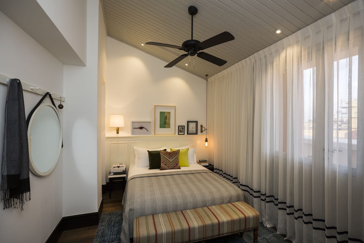 Une chambre à coucher comprenant un lit, un banc, un ventilateur de plafond, une table de chevet, une lampe, un miroir et des rideaux en voile.