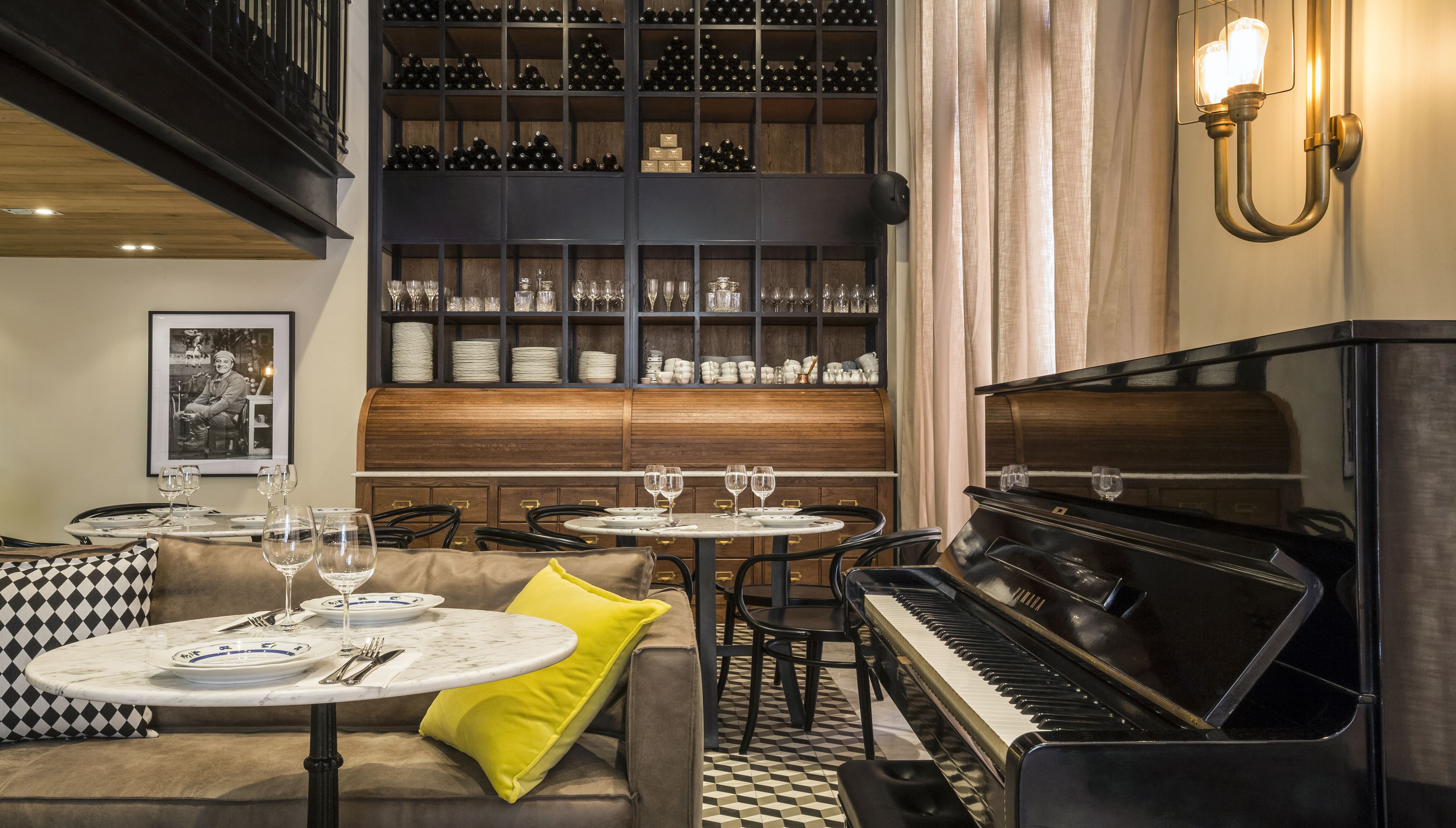 Un comedor con un piano, mesas y sillas. Hay botellas de vino y copas en estanterías.
