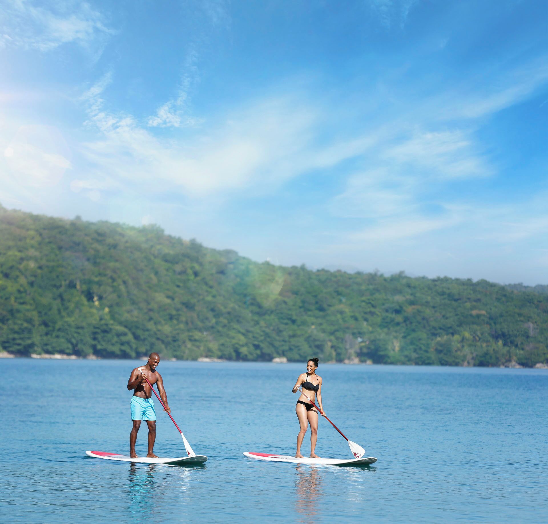 Dos personas haciendo paddleboard sobre un agua tranquila con una colina boscosa al fondo.