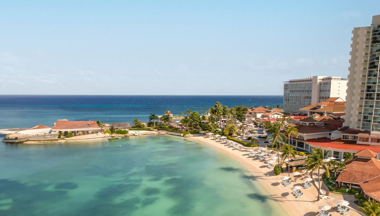 Un resort costero con playa, palmeras y un gran edificio de hotel.