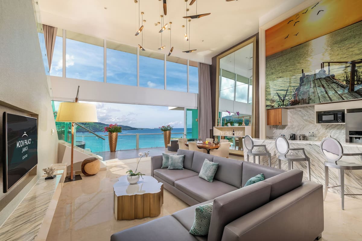 A modern living room with a sofa, coffee table, and a TV. There is a large window with a view of the sea.